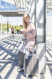 Spanien, Barcelona, junge Frau mit Trolley-Tasche und Mobiltelefon wartet am Bahnhof - GIOF05468