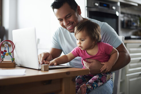 Smiling father and baby girl using laptop on table at home - ABIF01086