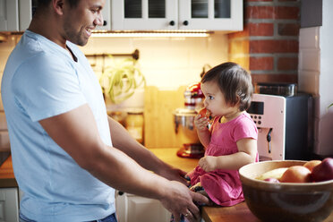 Smiling father looking at baby girl eating a tangerine on counter in kitchen at home - ABIF01094