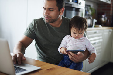 Father using laptop with baby girl using smartphone at home - ABIF01120