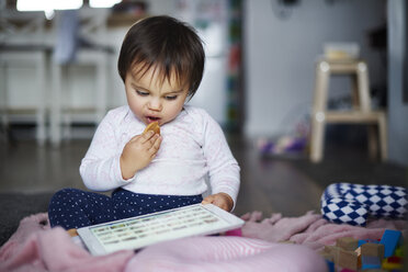 Baby girl sitting on the floor at home eating a cookie and using tablet - ABIF01123