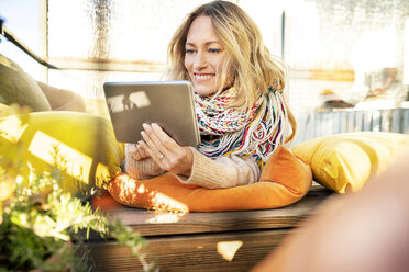 Porträt einer lächelnden blonden reifen Frau, die sich im Wintergarten mit einem digitalen Tablet entspannt - DMOF00095