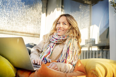 Porträt einer lächelnden blonden reifen Frau, die sich im Wintergarten am Laptop entspannt - DMOF00096