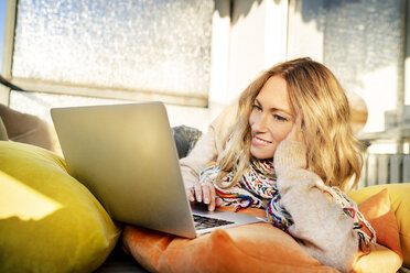 Porträt einer lächelnden blonden reifen Frau, die sich im Wintergarten am Laptop entspannt - DMOF00097