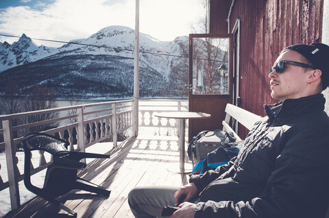 Mann sitzt auf einem Balkon mit schneebedeckten Bergen im Hintergrund in Troms Fylke, Norwegen, lizenzfreies Stockfoto