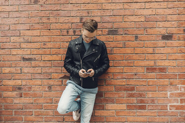 Teenage boy leaning on a brick wall looks at his phone in Sweden - FOLF09894