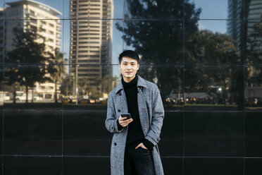Spain, Barcelona, portrait of young man with cell phone wearing black turtleneck pullover and grey coat - JRFF02444