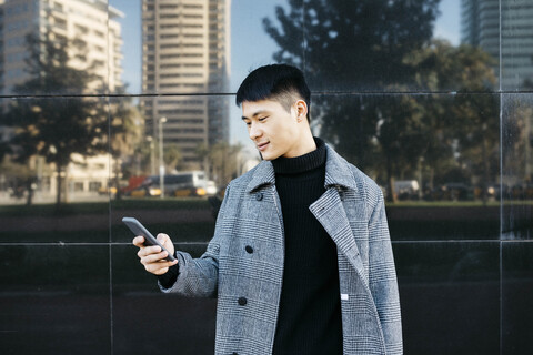 Spanien, Barcelona, junger Mann mit schwarzem Rollkragenpullover und grauem Mantel schaut auf sein Handy, lizenzfreies Stockfoto