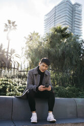 Spain, Barcelona, portrait of young man with mobile phone sitting on a wall - JRFF02448