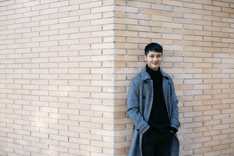 Porträt eines lächelnden jungen Mannes in grauem Mantel und schwarzem Rollkragenpullover, der an einer Wand lehnt, lizenzfreies Stockfoto