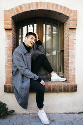 Laughing young man wearing fashionable coat sitting on window sill outdoors - JRFF02480