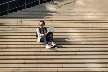 Lachender Mann mit Rucksack und Skateboard, der auf einer Treppe sitzt und einen Laptop benutzt - VABF02083