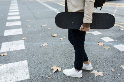 Mann mit Rucksack und Skateboard beim Überqueren der Straße im Herbst, Teilansicht, lizenzfreies Stockfoto