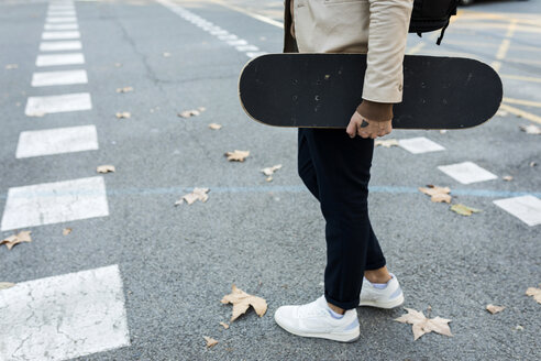 Mann mit Rucksack und Skateboard beim Überqueren der Straße im Herbst, Teilansicht - VABF02091