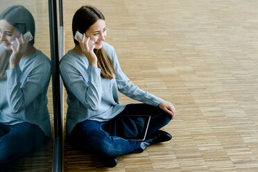 Frau sitzt im Schneidersitz an einer Glaswand und benutzt ein Mobiltelefon - CUF46635