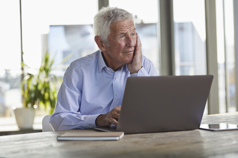 Portrait of pensive senior man sitting at table with laptop looking at distance stock photo