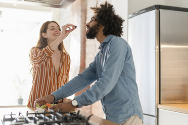 Couple standing in kitchen, preparing dinner party, woman feeding man with olive - ERRF00530