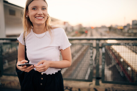 Porträt einer lächelnden jungen Frau, die ein Smartphone in der Hand hält, während sie auf einer Brücke in der Stadt steht, lizenzfreies Stockfoto