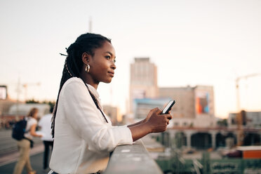 Thoughtful young woman looking away while holding mobile phone on bridge in city - MASF10300