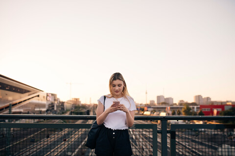 Junge Frau, die ein Mobiltelefon benutzt, während sie auf einer Brücke gegen den klaren Himmel in der Stadt steht, lizenzfreies Stockfoto