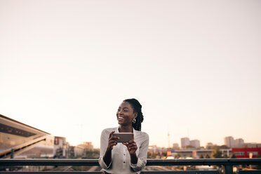 Lächelnde junge Frau, die wegschaut, während sie ein Mobiltelefon gegen den klaren Himmel in der Stadt hält - MASF10309