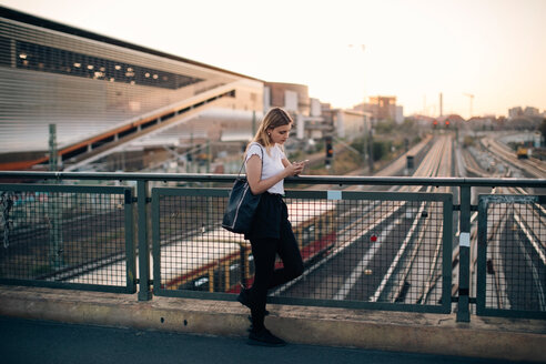 Junge Frau, die ein Mobiltelefon benutzt, während sie sich bei Sonnenuntergang auf einem Brückengeländer in der Stadt abstützt - MASF10312