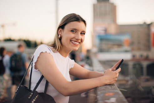 Porträt einer lächelnden jungen Frau, die ihr Handy in der Hand hält, während sie sich auf das Geländer einer Brücke in der Stadt stützt - MASF10314