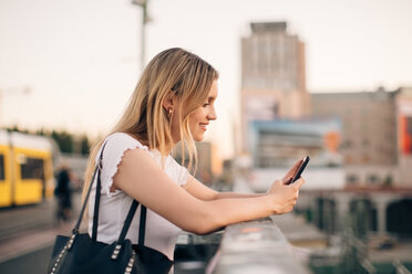 Seitenansicht einer jungen Frau, die ein Mobiltelefon benutzt, während sie sich am Geländer einer Brücke in der Stadt anlehnt - MASF10315