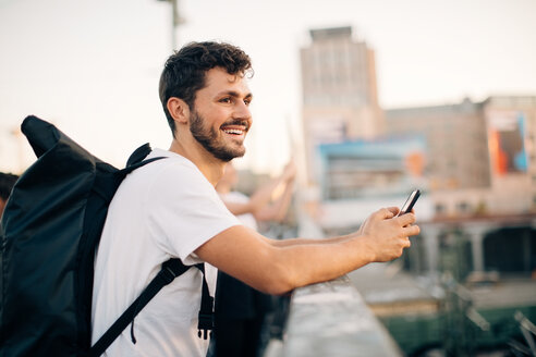 Seitenansicht eines lächelnden jungen Mannes, der wegschaut, während er ein Mobiltelefon an einer Brücke hält - MASF10318