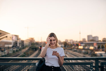 Lächelnde junge Frau mit Smartphone auf einer Brücke gegen den klaren Himmel bei Sonnenuntergang in der Stadt - MASF10319