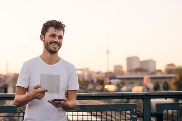 Smiling young man looking away while holding mobile phone on bridge against clear sky during sunset - MASF10320