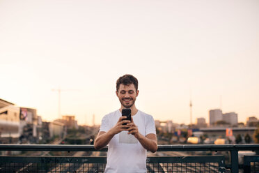Lächelnder junger Mann, der ein Mobiltelefon benutzt, während er auf einer Brücke gegen den klaren Himmel bei Sonnenuntergang steht - MASF10321