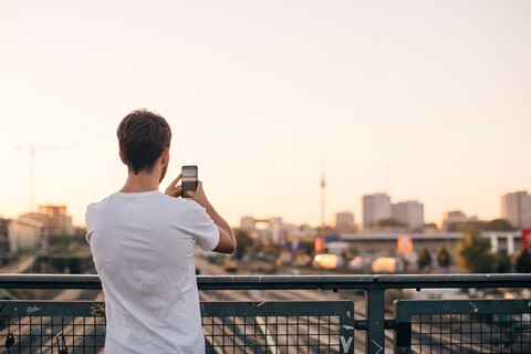 Rückansicht eines jungen Mannes, der auf einer Brücke stehend mit seinem Mobiltelefon die Stadt gegen den klaren Himmel fotografiert, lizenzfreies Stockfoto