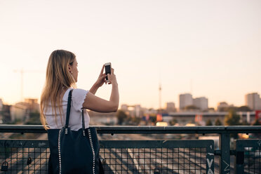 Junge Frau, die mit ihrem Handy fotografiert, während sie auf einer Brücke gegen den klaren Himmel bei Sonnenuntergang steht - MASF10323