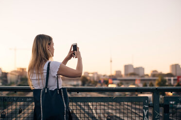 Young woman photographing through mobile phone while standing on bridge against clear sky during sunset - MASF10323