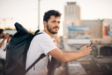 Seitenansicht eines jungen Mannes, der ein Mobiltelefon hält und sich an ein Brückengeländer lehnt - MASF10324