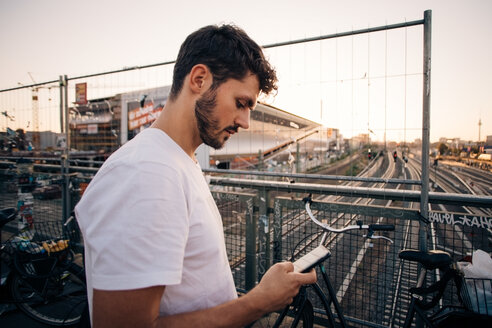 Seitenansicht eines jungen Mannes, der ein Mobiltelefon benutzt, während er auf einer Brücke über Bahngleise in der Stadt steht - MASF10327