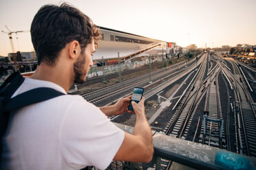 Young man using mobile phone while leaning on railing over railroad tracks in city - MASF10328