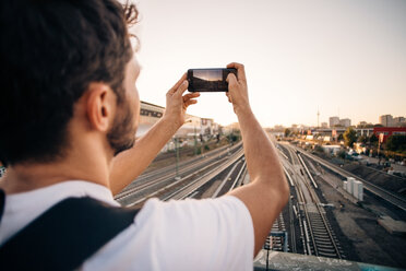 Young man photographing through mobile phone over railroad tracks in city - MASF10329