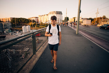 Junger Mann in voller Länge, der ein Mobiltelefon benutzt, während er auf dem Fußweg an einer Brücke in der Stadt geht - MASF10330