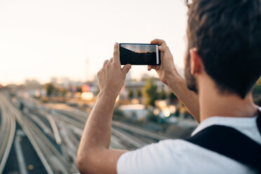 Young man photographing through smart phone over railroad tracks in city - MASF10331