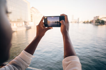 Cropped image of young woman photographing river through mobile phone in city - MASF10334