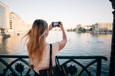 Rear view of young woman photographing river through mobile phone in city - MASF10337