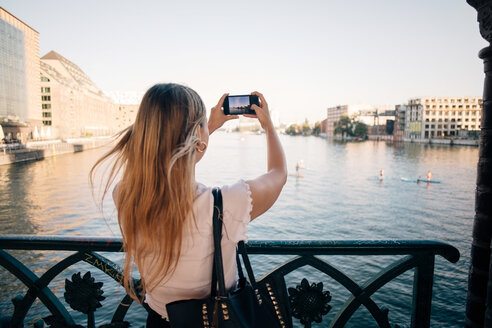 Rückansicht einer jungen Frau, die mit ihrem Handy einen Fluss in der Stadt fotografiert - MASF10337