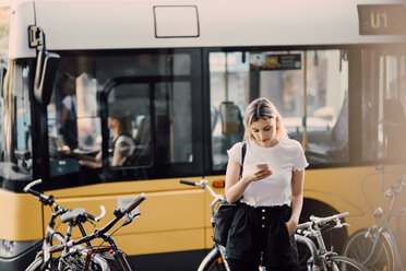 Young woman using smart phone while standing by bicycles against bus in city - MASF10339