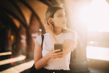 Young woman looking away while holding mobile phone on footpath in city during sunny day - MASF10341