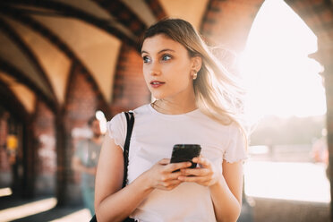 Young woman looking away while holding smart phone on footpath in city - MASF10344