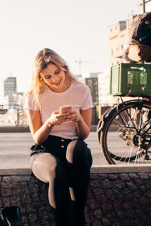 Lächelnde junge Frau, die ein Mobiltelefon benutzt, während sie auf einer Stützmauer an einer Straße in der Stadt sitzt - MASF10349