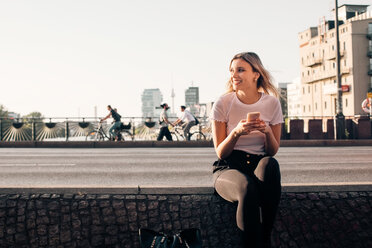 Smiling young woman holding mobile phone while sitting on retaining wall at street - MASF10350