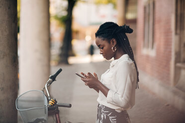 Side view of young woman using mobile phone while standing by bicycle on footpath in city - MASF10353